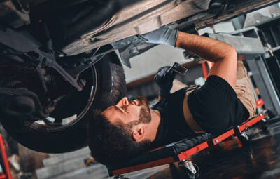 Bearded male technician in gloves lying on car creeper and using LED inspection lamp while working under automobile in garage. Bearded male technician in gloves lying on car creeper and using LED inspection lamp while working under automobile in garage. Side view. Close up view.